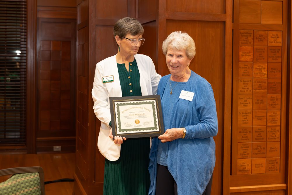Two individuals stand together, one holding a framed certificate, in a room with wooden paneling and a plaque wall.