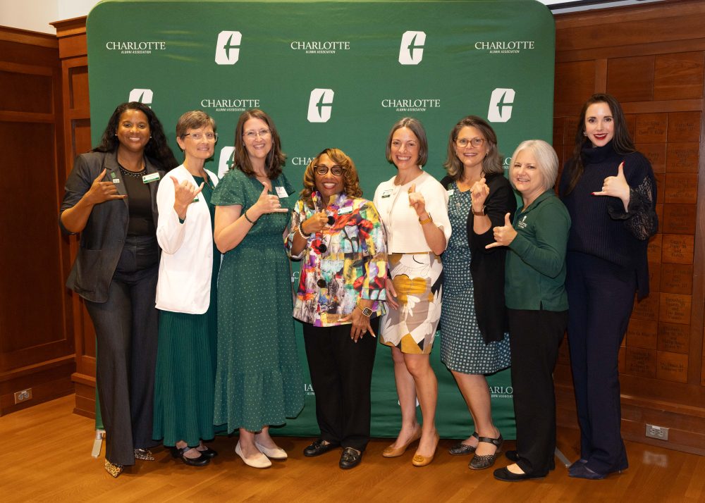Seven individuals pose together in front of a green backdrop featuring the Charlotte Alumni Association logo, making hand gestures.
