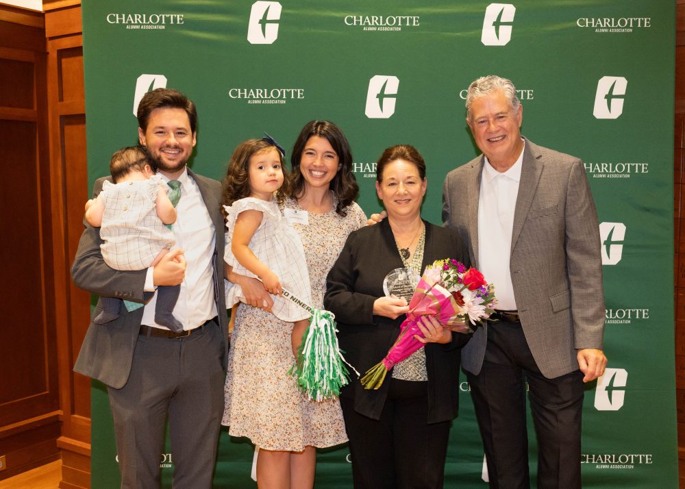 A family of five poses with a trophy and flowers in front of a green backdrop featuring the Charlotte Alumni Association logo.
