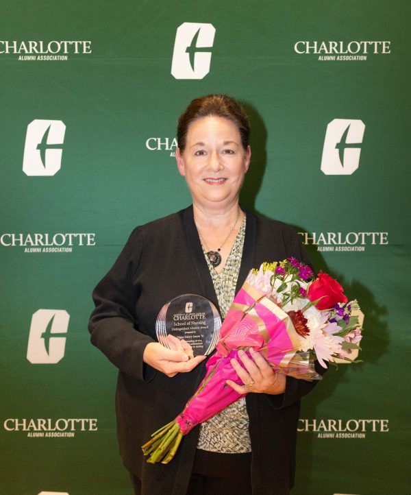 A person stands in front of a green backdrop with the Charlotte Alumni Association logo, holding a transparent award and a bouquet of flowers.