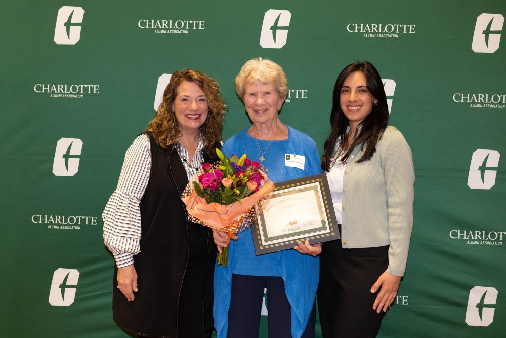 Three individuals pose together at a Charlotte Alumni Association event, with one holding a framed certificate and another holding a bouquet of flowers.